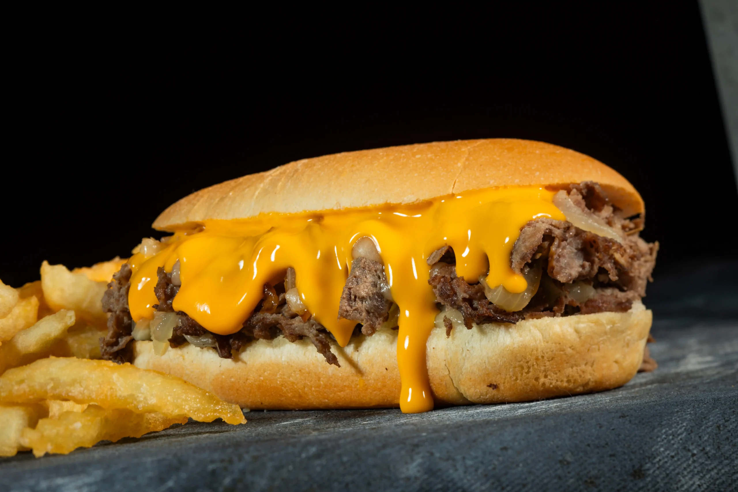 Philly cheesesteak sandwich with chopped beef, sautéed onions, and melted cheese sauce dripping over a toasted hoagie roll, served with fries